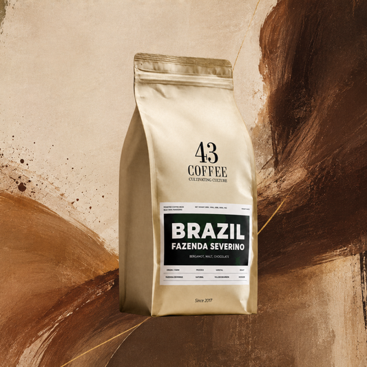 A bag of 43 Coffee Brazil Fazenda Severino whole bean coffee stands on a light surface with scattered coffee beans in front, set against an abstract warm brown and beige background.