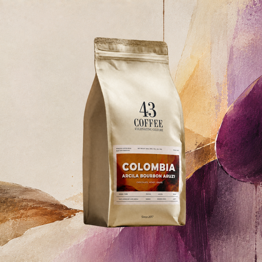 A stand-up pouch of 43 Coffee Colombia Arcila Bourbon Aruzzi coffee beans, featuring a metallic gold bag with a brown and orange label, set against an abstract painted background.